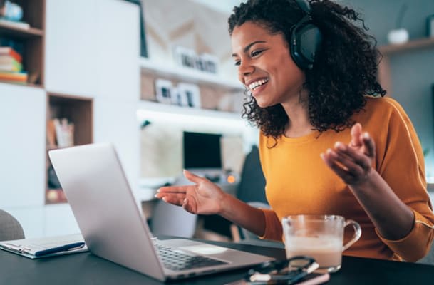 Une femme portant un casque audio est assise à son bureau, une tasse de café à portée de main. Elle sourit vers l'écran de son ordinateur en levant les mains.